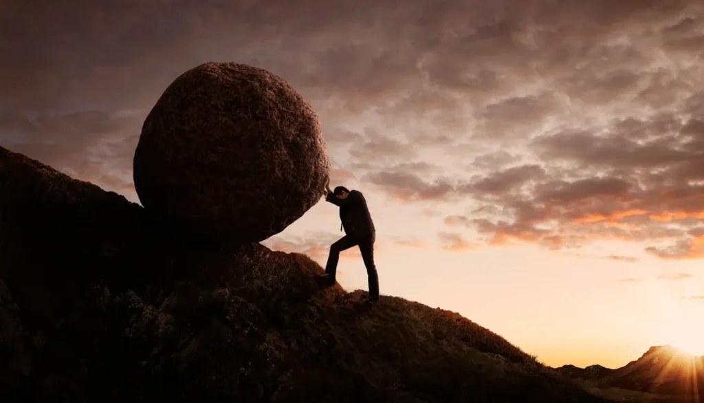 Person pushing large boulder uphill at sunset.